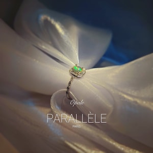 Op de afbeelding: Een zilverkleurige ring met een centrale ovale opaal omringd door een halo van kleine heldere steentjes. De ringband heeft een delicaat, getextureerd ontwerp. De tekst "Opale PARALLÈLE PARIS" staat in het wit onder de ring.