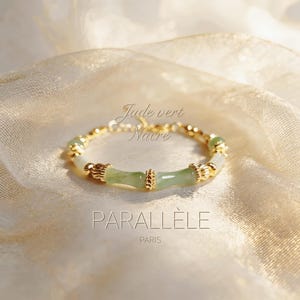 May include: A gold bracelet with jade and pearl accents. The bracelet features a central bamboo-style design in light green jade, with gold beads and accents. The text "Jade vert Nacre" and "PARALLÈLE PARIS" are visible.