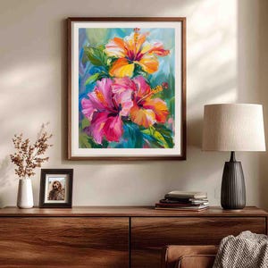 May include: A framed floral painting featuring vibrant hibiscus flowers in shades of pink, orange, and yellow. The artwork is set against a backdrop of blue and green hues, with a dark brown wooden frame. The painting is displayed on a wooden cabinet.