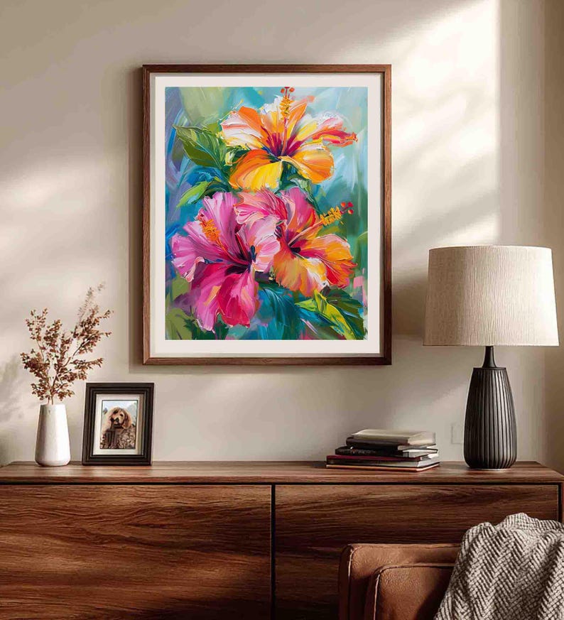 May include: A framed floral painting featuring vibrant hibiscus flowers in shades of pink, orange, and yellow. The artwork is set against a backdrop of blue and green hues, with a dark brown wooden frame. The painting is displayed on a wooden cabinet.
