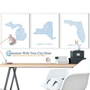 Może przedstawiać: Trzy akwarelowe wydruki stanów Michigan, Nowy Jork i Floryda. Każdy wydruk przedstawia zarys stanu w kolorze niebieskim i zawiera nazwę stanu oraz współrzędne. Wydruki są wyświetlane na białej ścianie nad biurkiem z lampą, krzesłem i innymi elementami dekoracyjnymi.