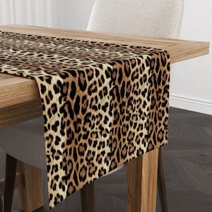 May include: A leopard print table runner on a wooden table. The runner has a repeating pattern of brown and black spots on a tan background. A neutral-colored chair is positioned behind the table.