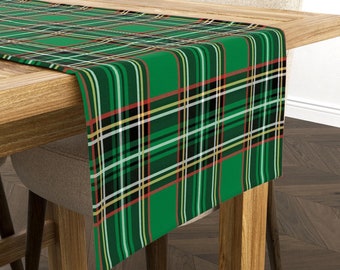 May include: A green tartan table runner featuring black, white, red, and yellow stripes. The runner is placed on a wooden table, with a neutral chair visible. The tartan design is a traditional pattern.
