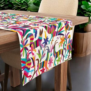 Peut inclure: Un chemin de table coloré avec un fond blanc orné d'un motif répétitif d'animaux stylisés, de fleurs et de formes géométriques. Le chemin de table est en tissu et a une bordure frangée.