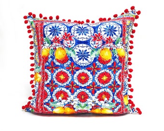 May include: A square cushion with a red, white and blue patterned design featuring lemons, flowers, and geometric shapes. The cushion has a red pom-pom trim.