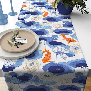 Puede incluir: Un camino de mesa blanco con un patrón floral azul y naranja con peces koi y libélulas. El camino de mesa está decorado con un patrón repetido de hojas de loto azules y flores blancas. El texto "HAVANA CONCEPT" está impreso en una pequeña tarjeta colocada en un plato beige.