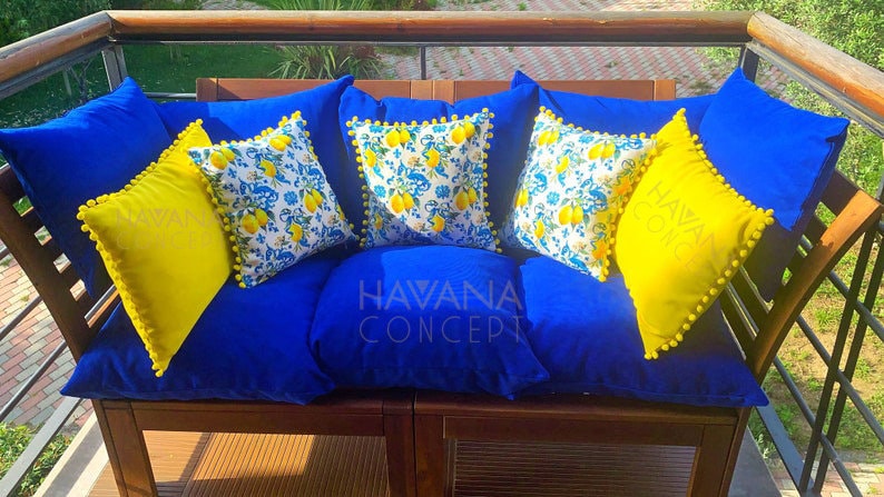 May include: A blue couch with yellow and white patterned throw pillows. The pillows have a pom-pom trim. The couch is on a wooden deck with a railing. The text "HAVANA CONCEPT" is visible on the couch.