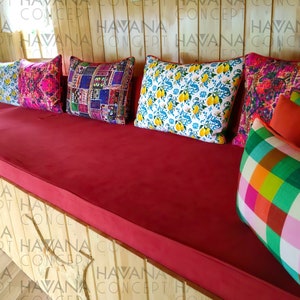 May include: A red cushioned bench with a variety of colorful patterned throw pillows. The pillows are arranged in a row on the bench, with the largest pillow in the center. The bench is made of wood and has a natural finish.