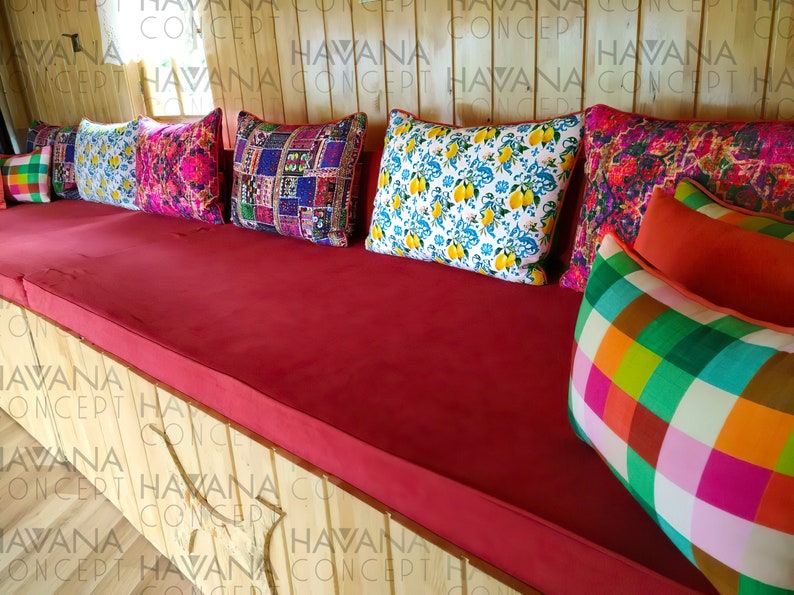 May include: A red cushioned bench with a variety of colorful patterned throw pillows. The pillows are arranged in a row on the bench, with the largest pillow in the center. The bench is made of wood and has a natural finish.