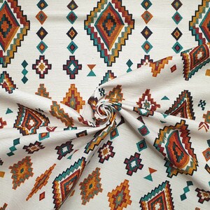 May include: White fabric with a repeating pattern of colourful geometric shapes. The pattern features a diamond shape with a smaller diamond inside, surrounded by smaller geometric shapes in shades of orange, red, teal, and brown.