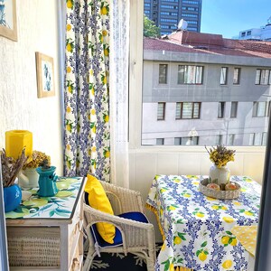 May include: A white wicker chair with a yellow pillow sits on a blue and black rug. A white table with a lemon print tablecloth is set with a vase of dried flowers and a wicker tray. The table is in front of a window with a view of a city.