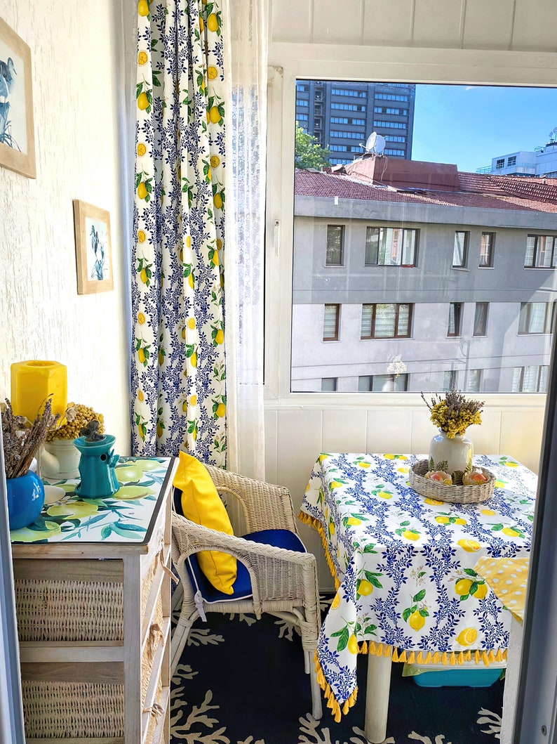 May include: A white wicker chair with a yellow pillow sits on a blue and black rug. A white table with a lemon print tablecloth is set with a vase of dried flowers and a wicker tray. The table is in front of a window with a view of a city.