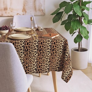 Puede incluir: Una mesa de comedor rectangular cubierta con un mantel de tela con un estampado de leopardo en tonos marrón, negro y beige. La mesa está puesta con platos blancos, copas de cristal transparente, cubiertos dorados y un cuaderno marrón cerrado con un bolígrafo.