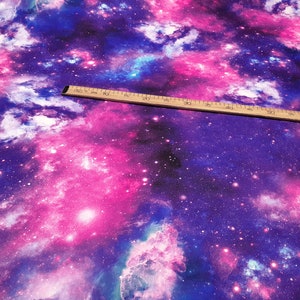 Könnte beinhalten: Stoff mit einem lebendigen Galaxiemuster in Lila, Rosa und Blau. Ein Holzlineal liegt auf dem Stoff. Der Stoff eignet sich für Bastel- oder Nähprojekte.