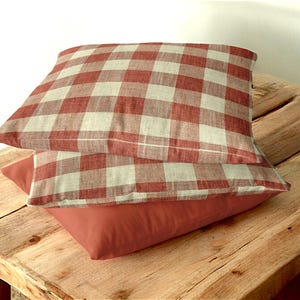 Puede incluir: Dos almohadas de tiro a cuadros rojos y blancos apiladas una encima de la otra sobre una superficie de madera.