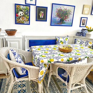 May include: White wicker dining chairs with blue cushions and a white tablecloth with a lemon print. The table is set with a wooden bowl and a blue and white rug is on the floor.
