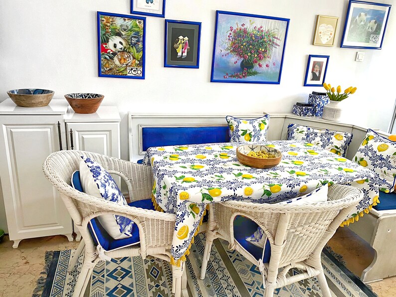 May include: White wicker dining chairs with blue cushions and a white tablecloth with a lemon print. The table is set with a wooden bowl and a blue and white rug is on the floor.