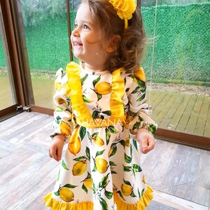 May include: A little girl wearing a white dress with yellow lemons and green leaves printed on it. The dress has yellow ruffles around the neckline and hem. She is wearing a yellow flower headband. The dress has the text "HAVANA" on the front.