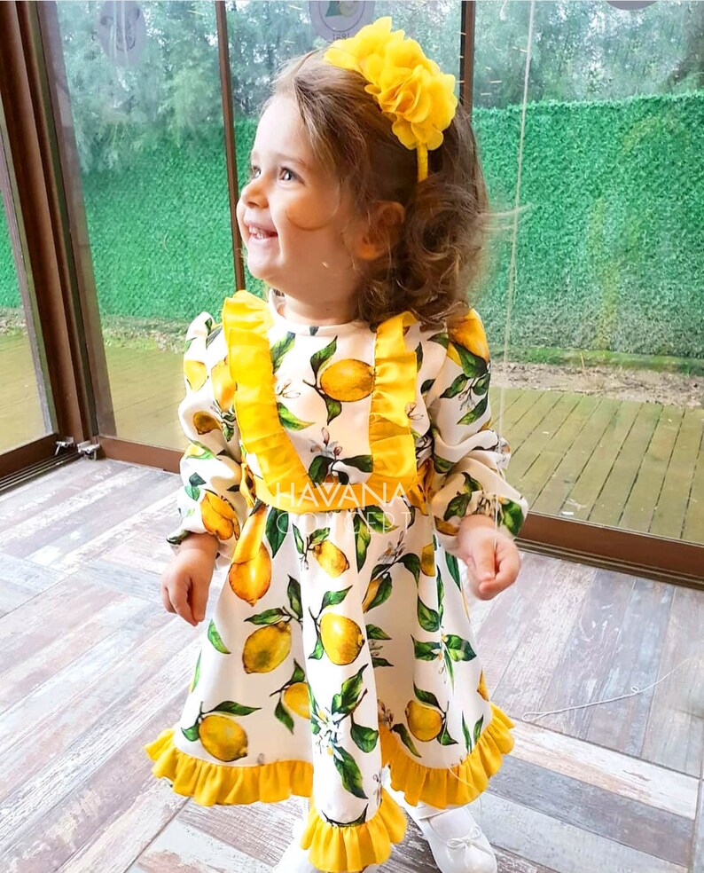 May include: A little girl wearing a white dress with yellow lemons and green leaves printed on it. The dress has yellow ruffles around the neckline and hem. She is wearing a yellow flower headband. The dress has the text "HAVANA" on the front.