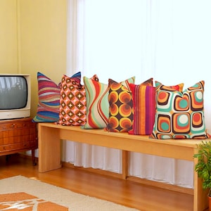 Puede incluir: Una colección de cojines decorativos con patrones vibrantes de inspiración retro. Los cojines presentan formas geométricas, líneas onduladas y diseños circulares en una variedad de colores, incluyendo naranja, turquesa y marrón. Los cojines están colocados en un banco de madera.