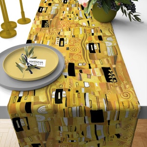 Puede incluir: Un camino de mesa amarillo con un patrón geométrico inspirado en la famosa pintura "El Beso" de Gustav Klimt. El camino de mesa presenta un patrón repetitivo de cuadrados, rectángulos y espirales en tonos dorados, negros y blancos.