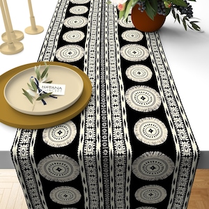 May include: Black and white table runner with a geometric pattern featuring circles and fringe. The runner has a repeating pattern of circles with a diamond shape in the center. The circles are surrounded by a border of small squares and diamonds. The runner is made of a soft, woven fabric.