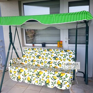 May include: A green striped canopy covers a white and yellow lemon patterned swing. The swing is made of metal and has a white cushion. The swing is on a patio with a white door and a tiled floor.