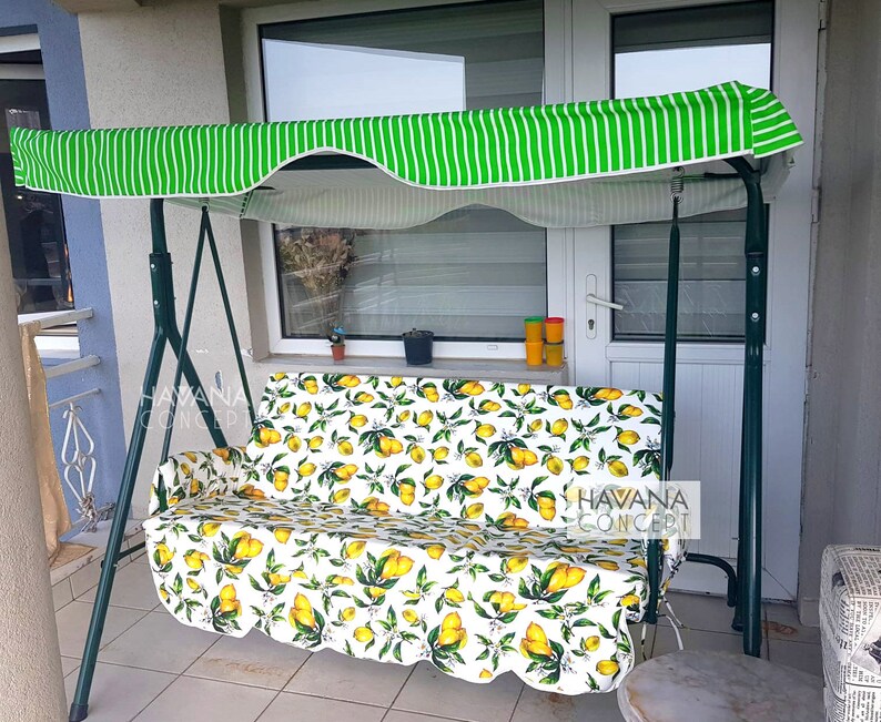 May include: A green striped canopy covers a white and yellow lemon patterned swing. The swing is made of metal and has a white cushion. The swing is on a patio with a white door and a tiled floor.