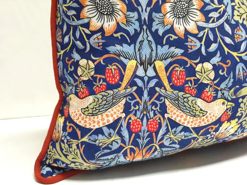 Pu&ograve; includere: Un cuscino blu con un bordo rosso presenta un motivo ripetitivo di uccelli stilizzati, fiori e foglie. Il motivo &egrave; ispirato al movimento Arts and Crafts.