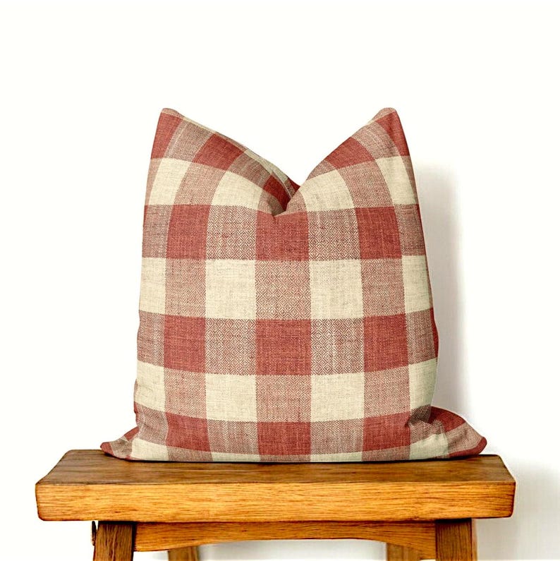 Puede incluir: Una almohada de cuadros rojos y blancos sobre un banco de madera. La almohada est&aacute; hecha de una tela suave y tejida con una textura sutil.