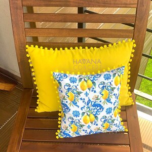 May include: Two pillows on a wooden chair. The bottom pillow is yellow with white pom-poms around the edge and the text "HAVANA CONCEPT" embroidered on it. The top pillow is white with a blue and yellow lemon print and white pom-poms around the edge.