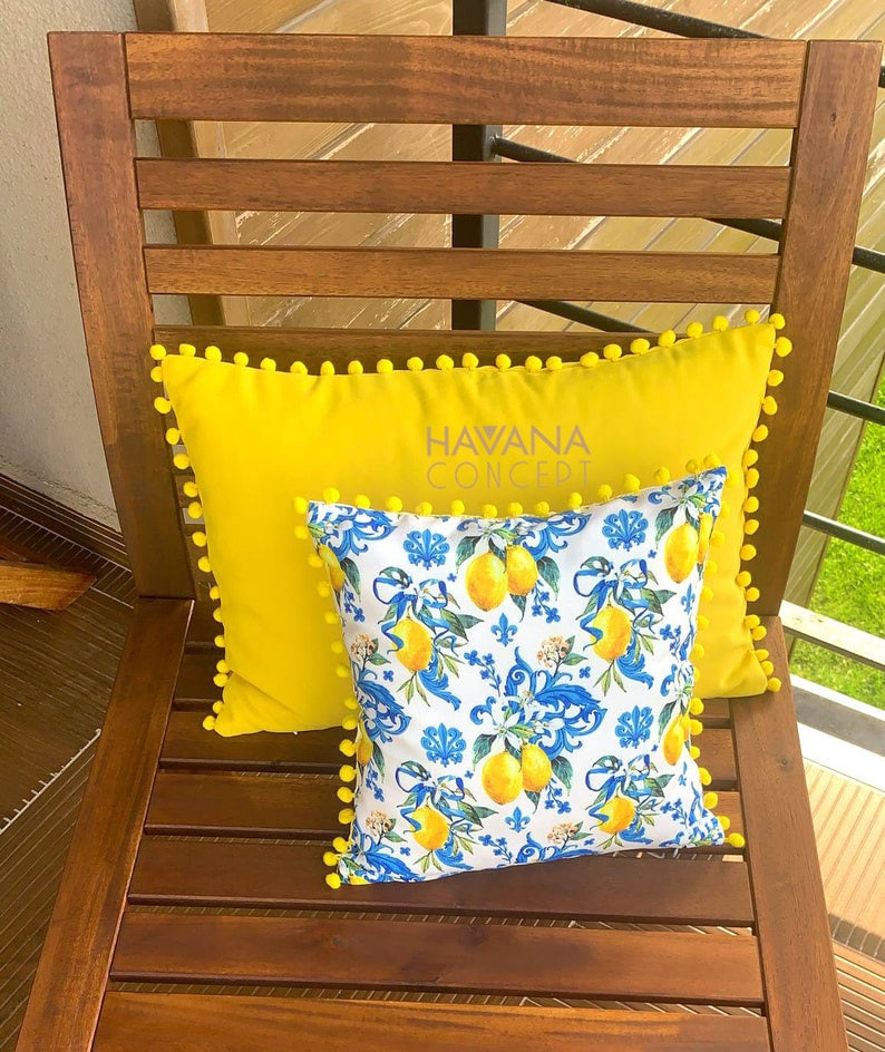 May include: Two pillows on a wooden chair. The bottom pillow is yellow with white pom-poms around the edge and the text "HAVANA CONCEPT" embroidered on it. The top pillow is white with a blue and yellow lemon print and white pom-poms around the edge.