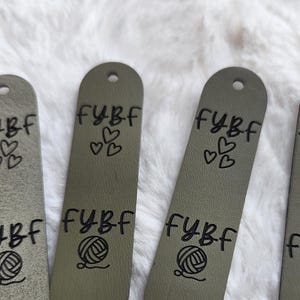 May include: Several olive green rectangular tags with rounded ends, each featuring a pre-punched hole at the top and bottom. The tags are printed with the black text FYBF, three small heart outlines, and a graphic of a ball of yarn.