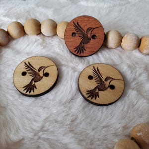 May include: Three wooden buttons featuring a hummingbird design. Two buttons are light brown and one button is dark brown.