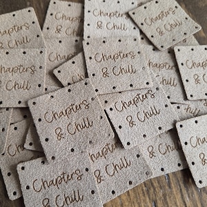 May include: A collection of square, light tan faux leather tags featuring the laser-engraved text "Chapters & Chill" in a cursive font. Each tag has pre-punched holes along the edges for sewing, with a textured surface and dark brown lettering.