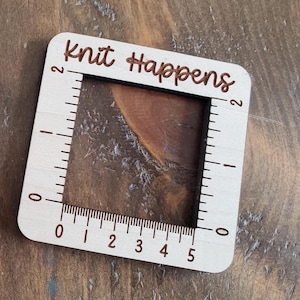 May include: A wooden ruler with a square cutout and the text "Knit Happens" on the top. The ruler has markings in inches from 0 to 5.
