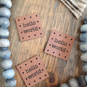 May include: Three brown leather patches with the text "hello world" in black lettering. The patches are on a wooden surface.