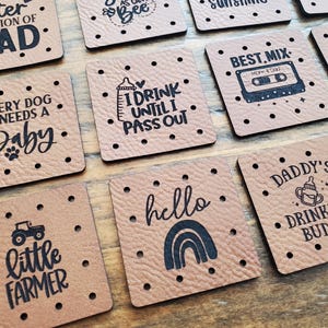May include: A collection of square tan faux leather patches with pre-punched holes along the edges. Each patch features black printed text and graphics, including phrases like Cuter Version of Dad, Every Dog Needs a Baby, I Drink Until I Pass Out, Best Mix, Little Farmer, Hello, and Daddy's Drinking Buddy.