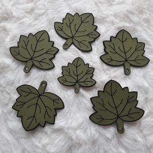May include: Six olive green leaf-shaped embellishments with black outlines and a small hole at the base. The leaves are arranged on a white, fluffy surface, showcasing their detailed veining.