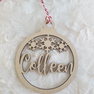 May include: A wooden Christmas ornament with the name "Colleen" in cursive script. The ornament has a circular design with snowflake cutouts and a red and white twine hanger. The ornament is made of light-colored wood.