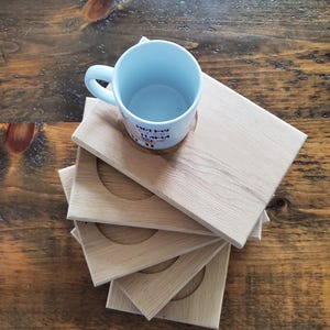 May include: A light blue ceramic mug with the text "Hot My Llama" rests on a wooden coaster. Several additional square wooden coasters with circular cutouts are stacked below. The coasters are made of light-colored wood.