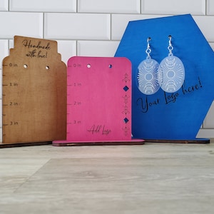 Peut inclure: Trois présentoirs à boucles d'oreilles en bois. Le présentoir de gauche est brun avec une règle graduée en pouces. Le présentoir du milieu est rose avec une règle graduée en pouces et le texte "Add Logo". Le présentoir de droite est bleu avec une forme hexagonale et le texte "Your Logo here!" Deux paires de boucles d'oreilles blanches sont exposées sur le présentoir bleu.