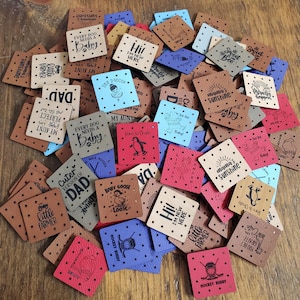 May include: A pile of small, square tags made to look like leather in shades of tan, red, blue, and brown. Each tag has a unique design and text, such as "Every Dog Needs a Baby" and "Hi! I'm New Here."