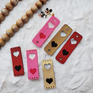 May include: Six leather heart-shaped cutouts in pink, red, and brown. Each cutout has a smaller heart in the center, either black or white.