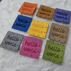 May include: A collection of nine square labels in various colours including orange, mustard, purple, blue, yellow, tan, grey, pink, and green. Each label features the text hello world embossed in the centre and small holes along the edges.