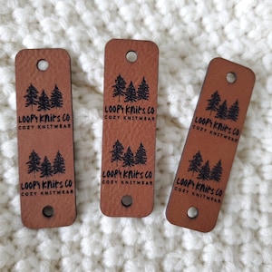 May include: Three rectangular brown faux leather tags with rounded corners, each featuring two pre-punched holes. The tags are imprinted with black text reading "LOOPY KNITS CO COZY KNITWEAR" and a graphic of three pine trees repeated twice.