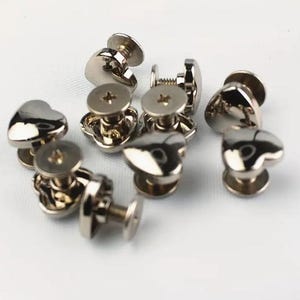 May include: A collection of shiny, silver heart-shaped rivets. Each rivet has a heart-shaped head and a screw-like base. The rivets are scattered on a white surface, reflecting light.