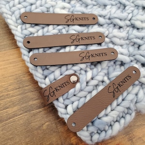 May include: Five brown rectangular labels with the text SG KNITS engraved in black. Four labels feature holes at each end for attachment, while one label is folded and secured with a silver-coloured metal rivet, resting on a light blue knit fabric.
