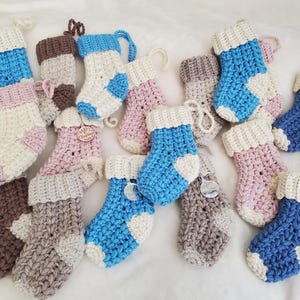 May include: A selection of miniature, crocheted Christmas stockings in shades of blue, pink, brown, and grey. Each stocking features a white cuff and heel, with a loop for hanging. Some have small wooden or metal charms attached.