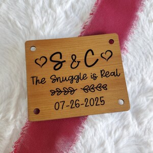 May include: A rectangular light brown wooden plaque with four corner holes, featuring black engraved text that reads S & C with heart icons, The Snuggle is Real, decorative leaf flourishes, and the date 26-07-2025 centred on the surface.